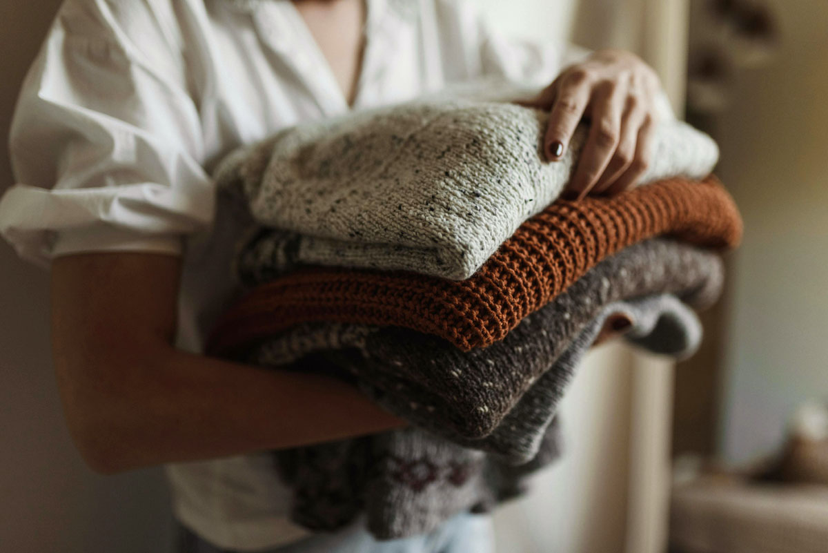 A woman in a white tshirt carries a pile of several folded brown and grey sweaters. 