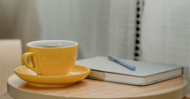 A grey gratitude journal lays on a wooden table next to a yellow cup of coffee.