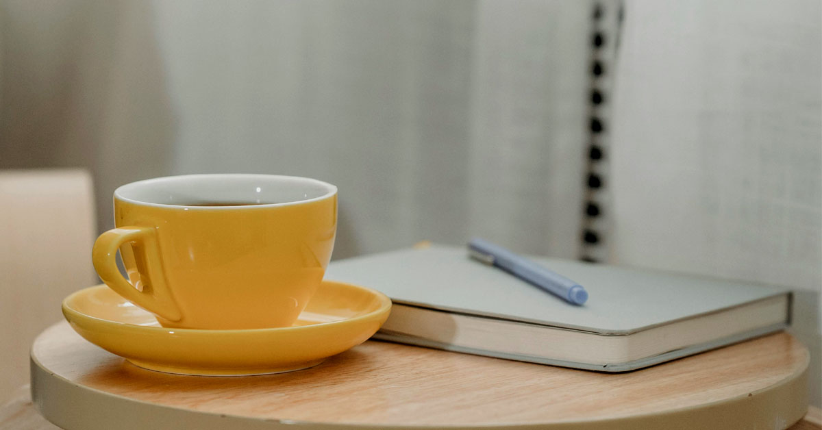 A grey gratitude journal lays on a wooden table next to a yellow cup of coffee.