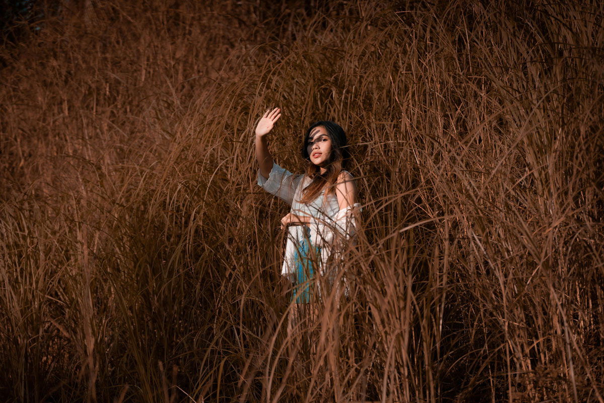 A woman stands in a field of wild bushes shading her eyes from the setting sun.