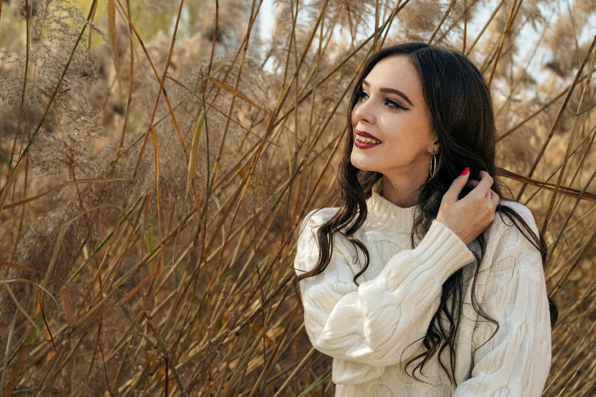 A woman with black hair and a white sweater stands smiling in a field of tall grass. 