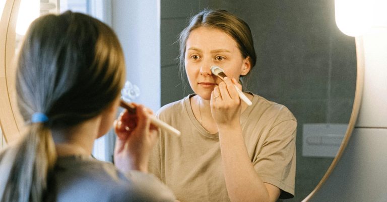 A woman looks at her reflection in a mirror while putting on makeup.
