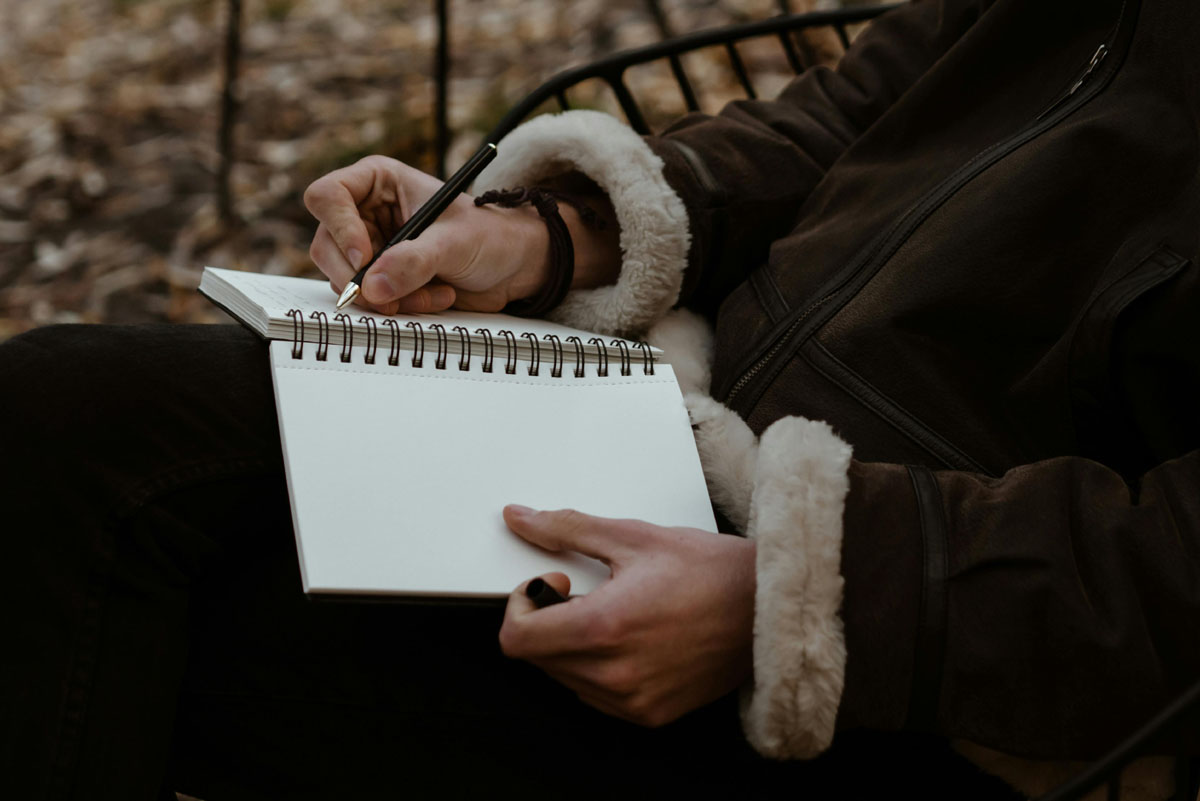 A woman writes in a spiral bound journal with autumn leaves in the background.