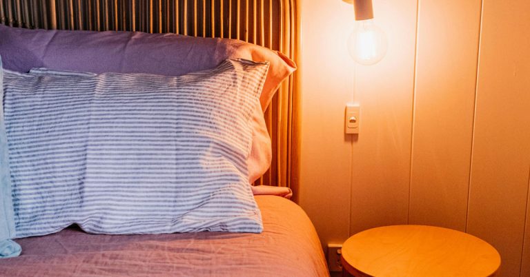 Pillows lay on a bed alongside a round nightstand bathed in a golden light that's an essential part of my evening routine.