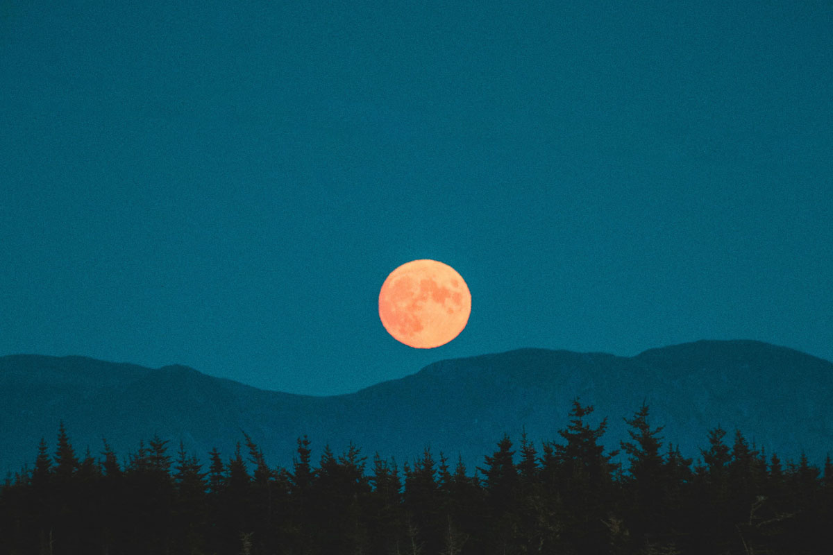 A full moon shines in a blue sky with mountains and trees in the background.