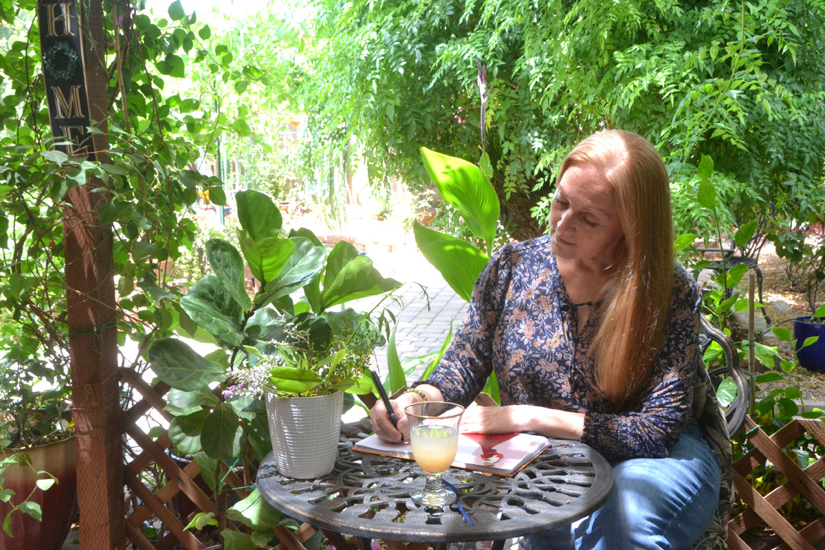 Dee sits under a tree in the garden writing june journal prompts in her notebook.