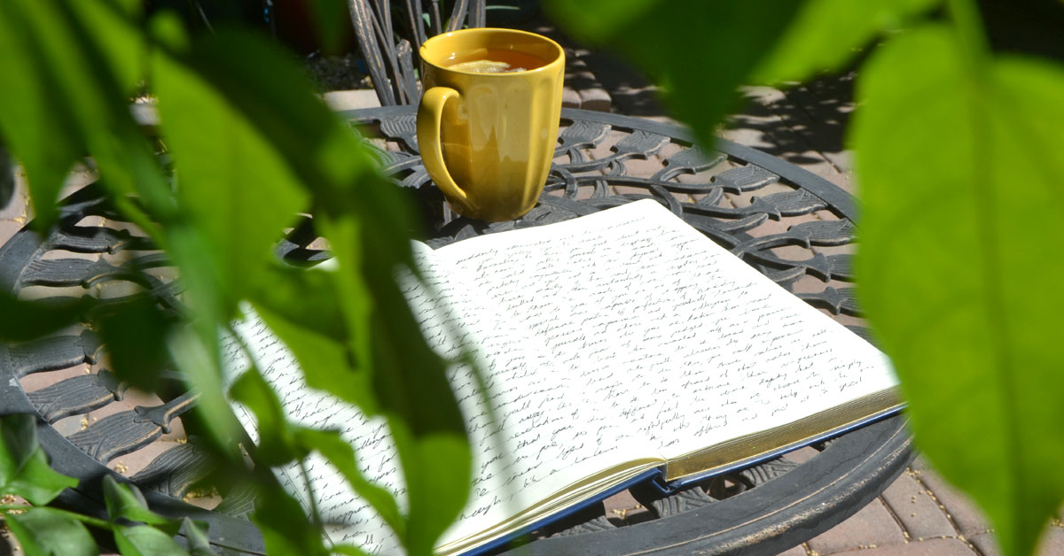 A journal lays open in the sun under a tree next to a yellow mug.