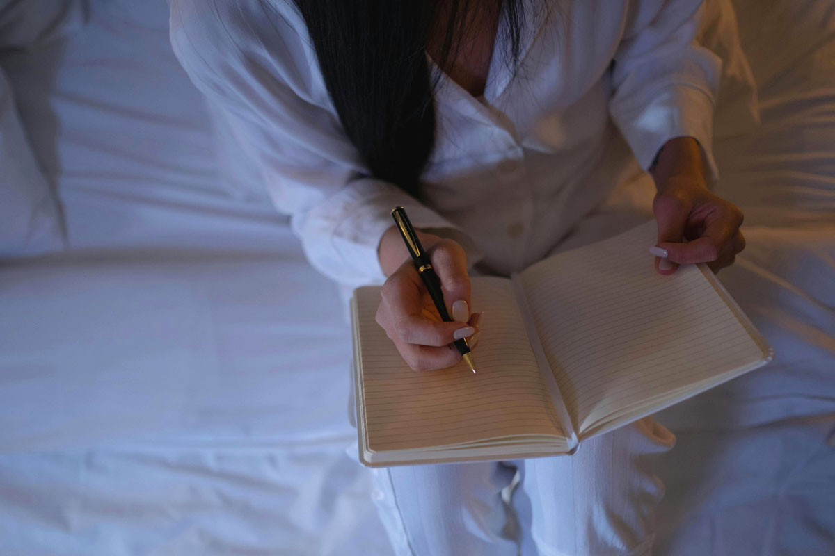 A woman journals in bed wearing white pajamas and sitting on white sheets.