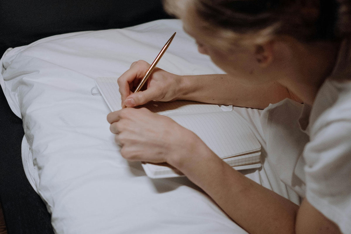 A woman lays on a pillow and writes in a journal.