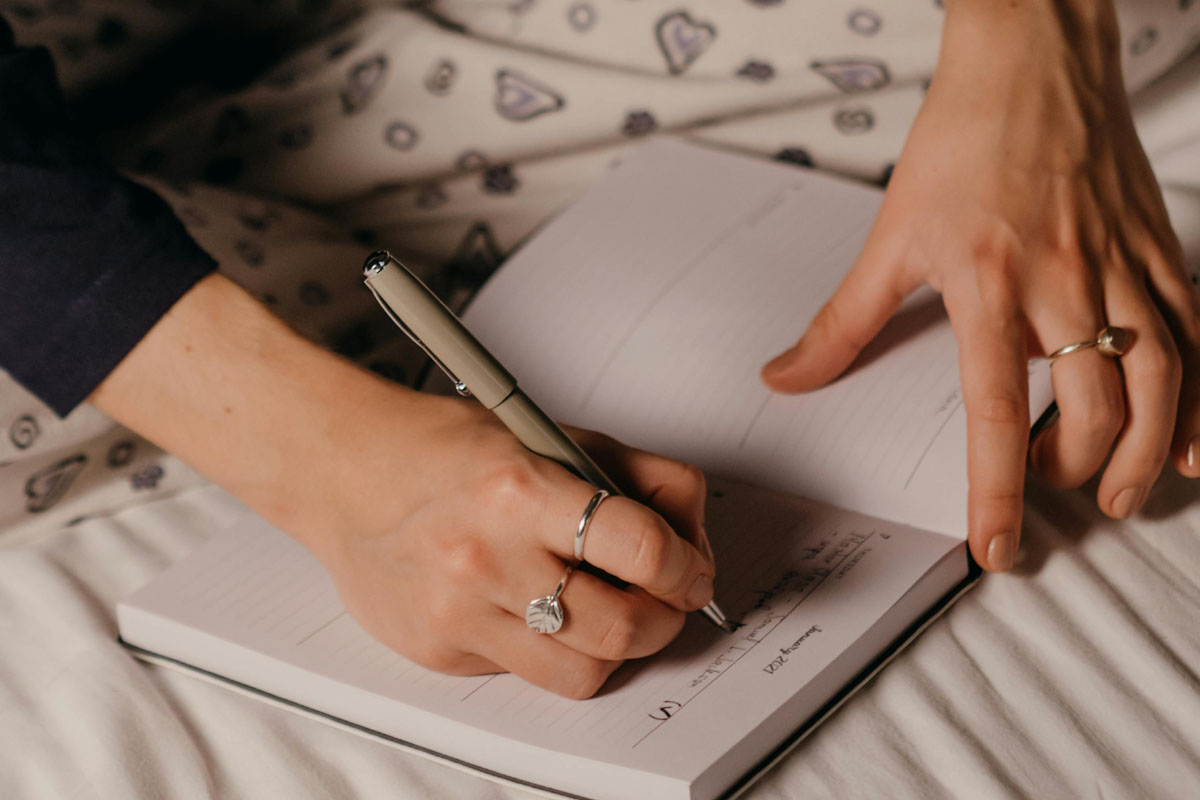 A woman writes in a journal in bed against some beige sheets.