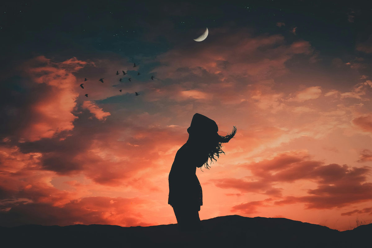 A woman stands against a setting sun with a crescent moon in the background as part of a new moon rituals.