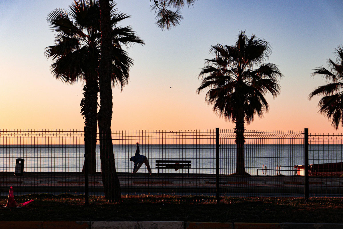 A woman does stretches on a beach boardwalk with a sunset and palms in the background.