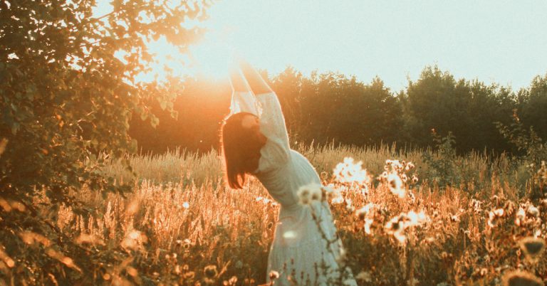 A woman in a field of tall greenery lifts her arms up to stretch against the setting sun.