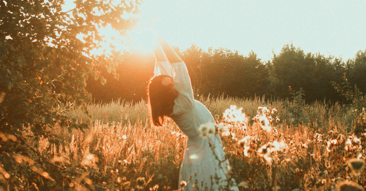 A woman in a field of tall greenery lifts her arms up to stretch against the setting sun.