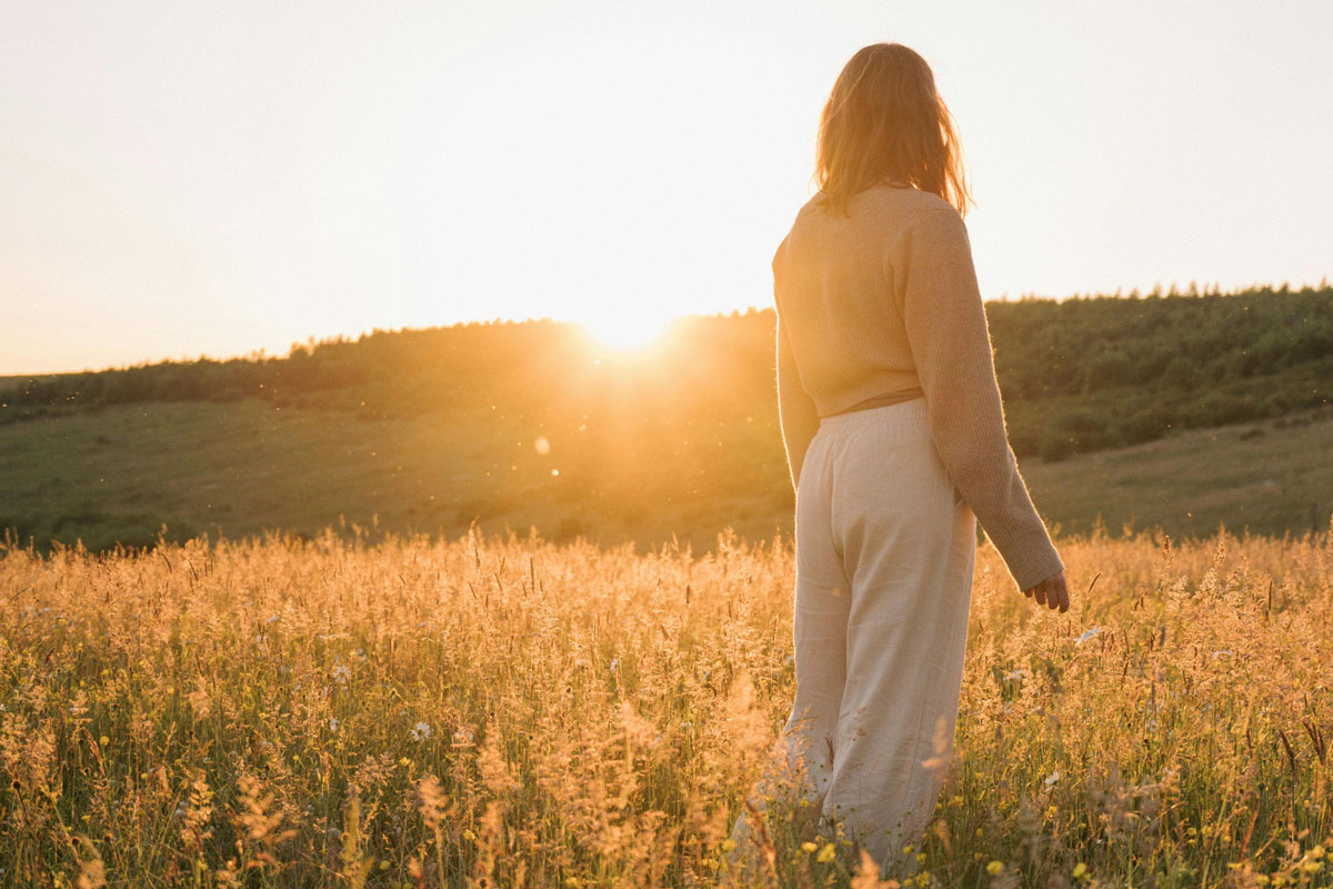 A woman stands in a golden field of grass as the sun sets.