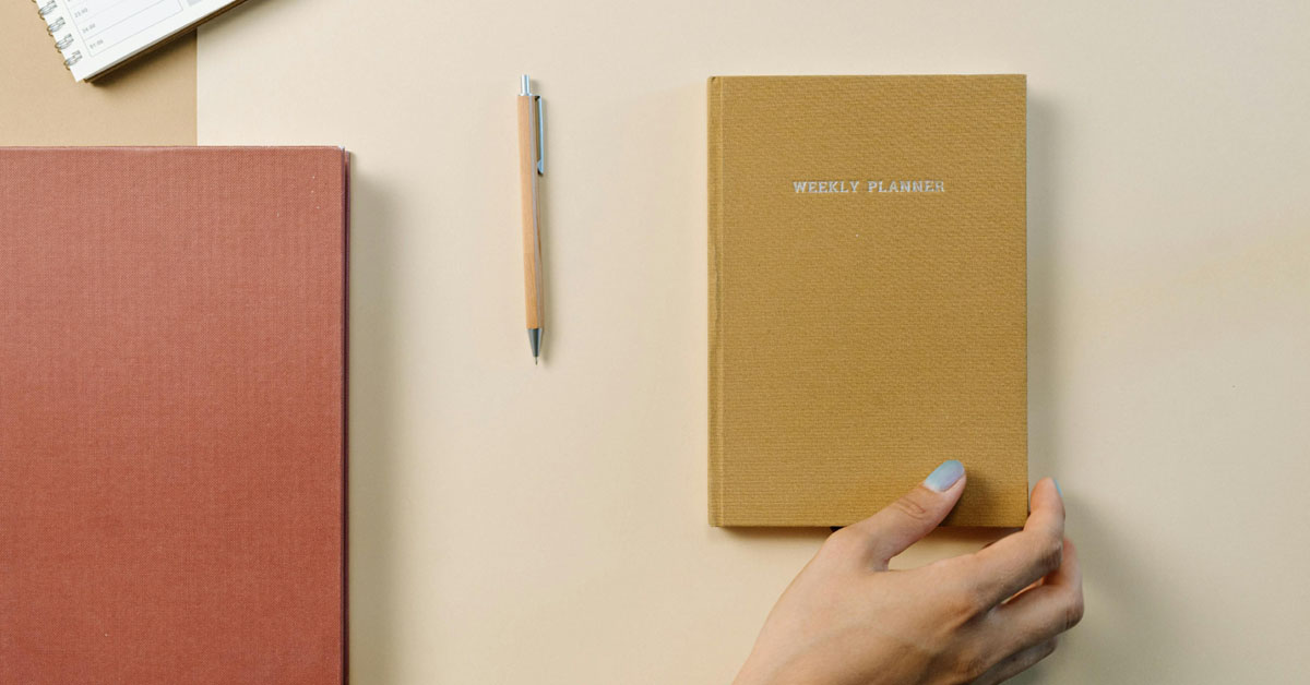 A hand reaches for a yellow ochre bullet journal laying on a beige desk alongside a pencil.