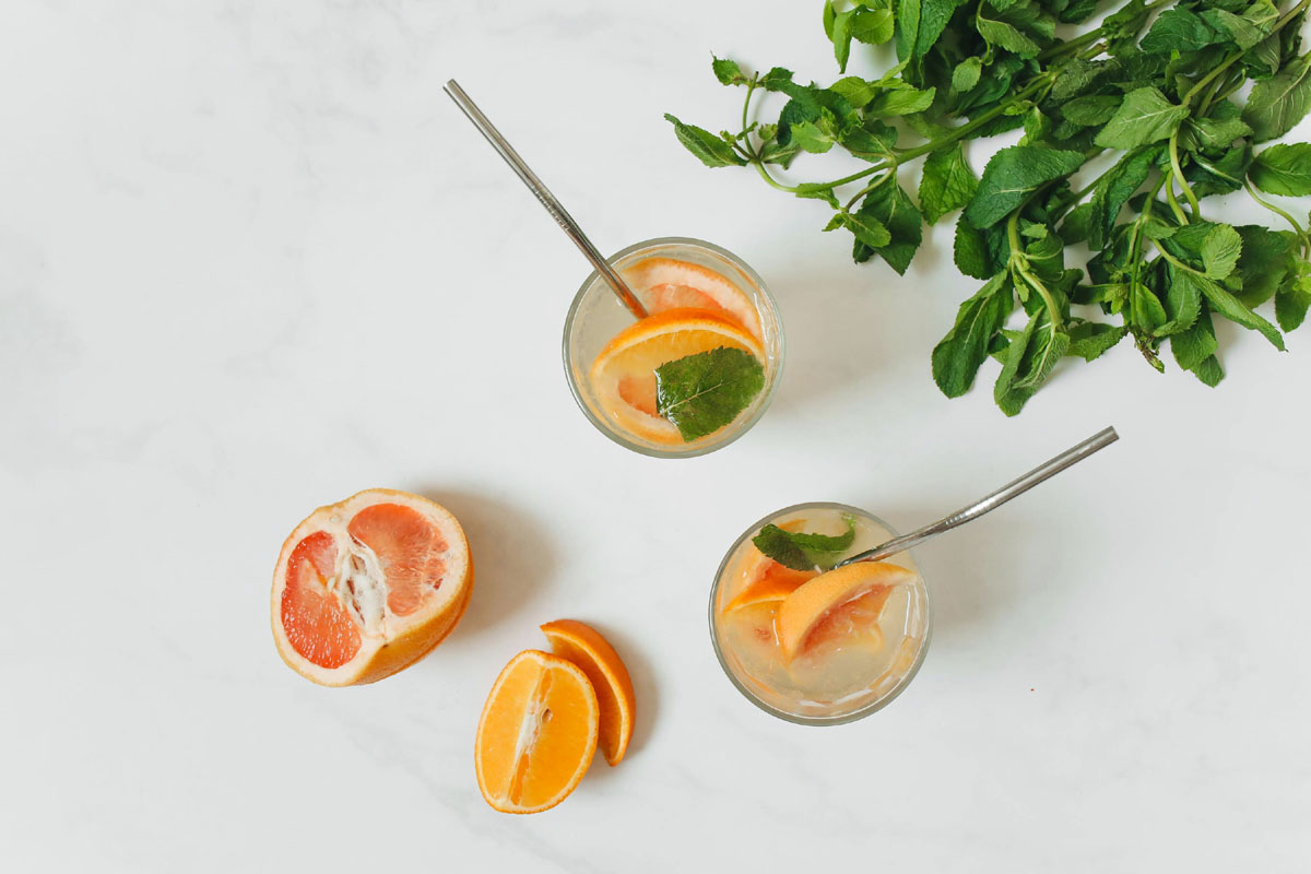 Two glasses of grapefruit juice stand on a white table with mint and fruit slices around.