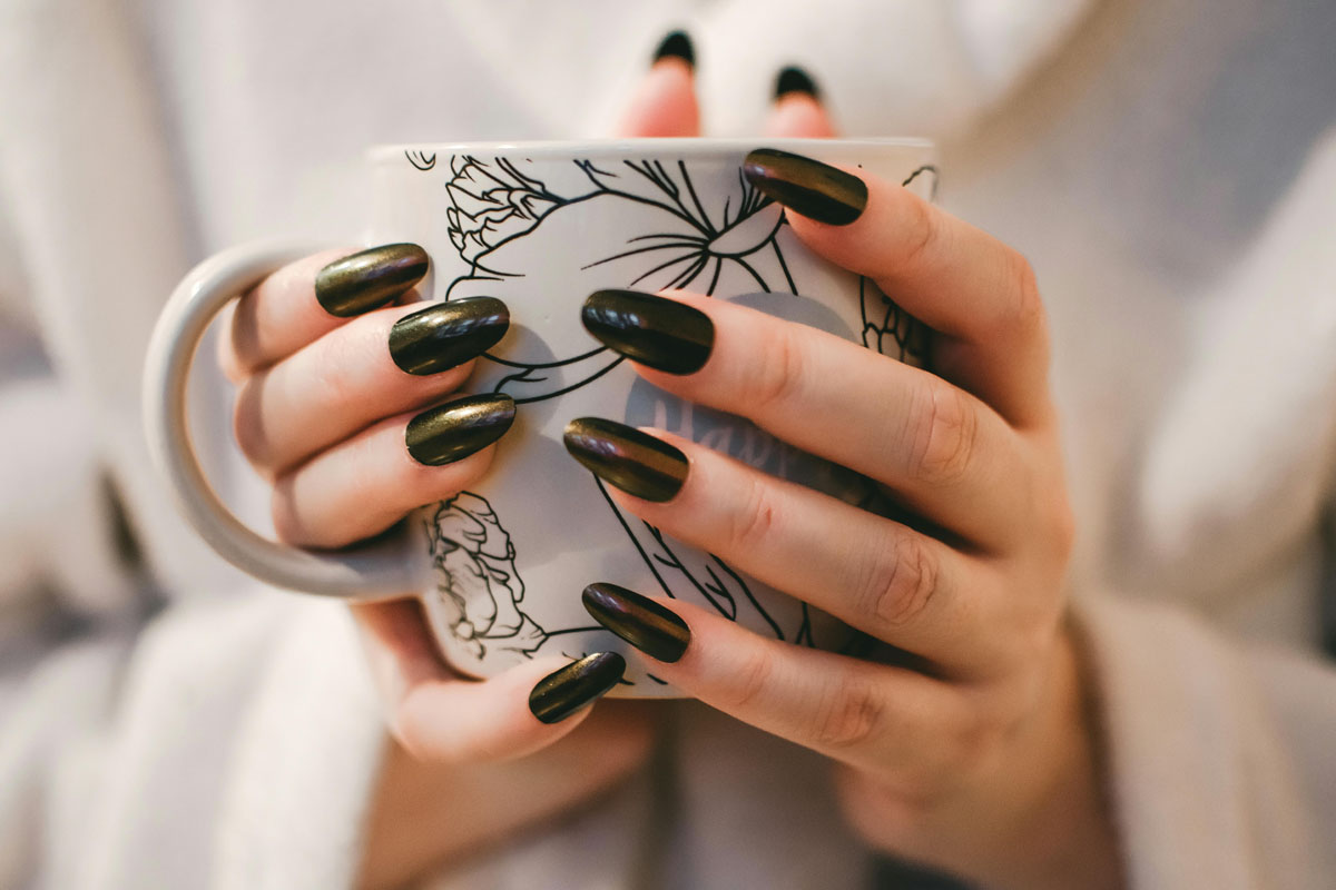 A woman with long black nails holds a cup of coffee in her hand.