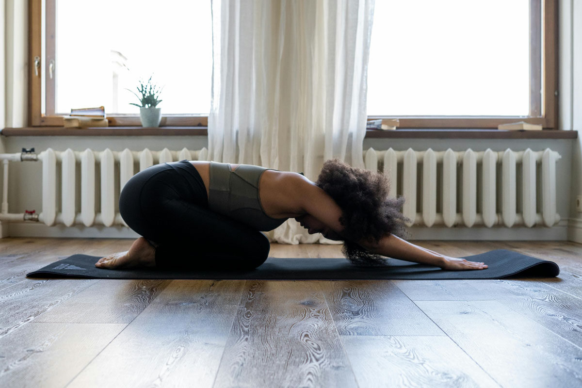 A woman does yoga stretches on the floor with wide windows behind her.