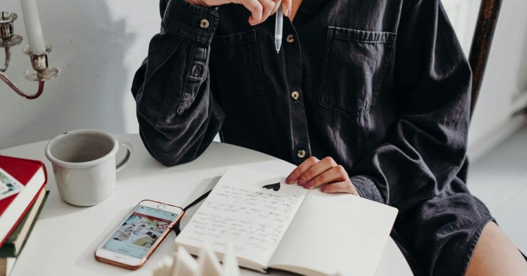 A woman in a dark shirt sits at a desk with an open notebook and phone in front of her as part of some creative morning routine ideas..