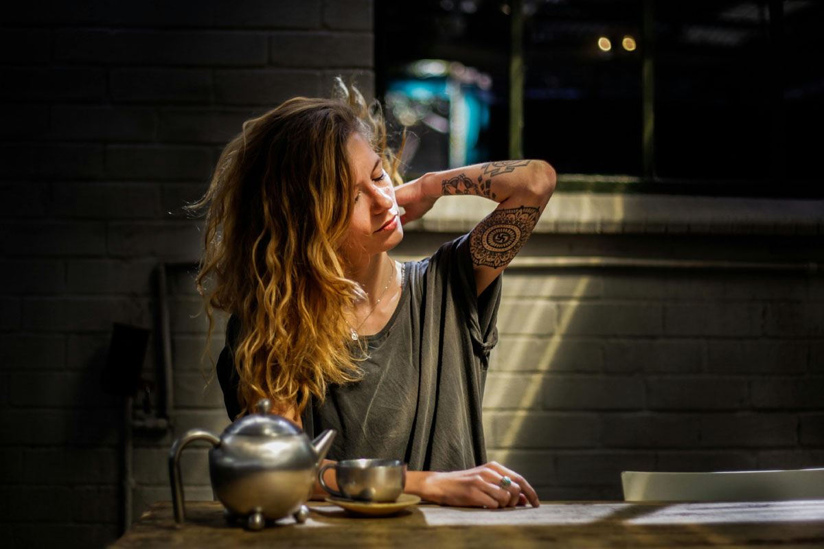 A woman with blond hair in a grey tshirt sits at a desk rubbing her shoulder with a kettle of tea in front of her.
