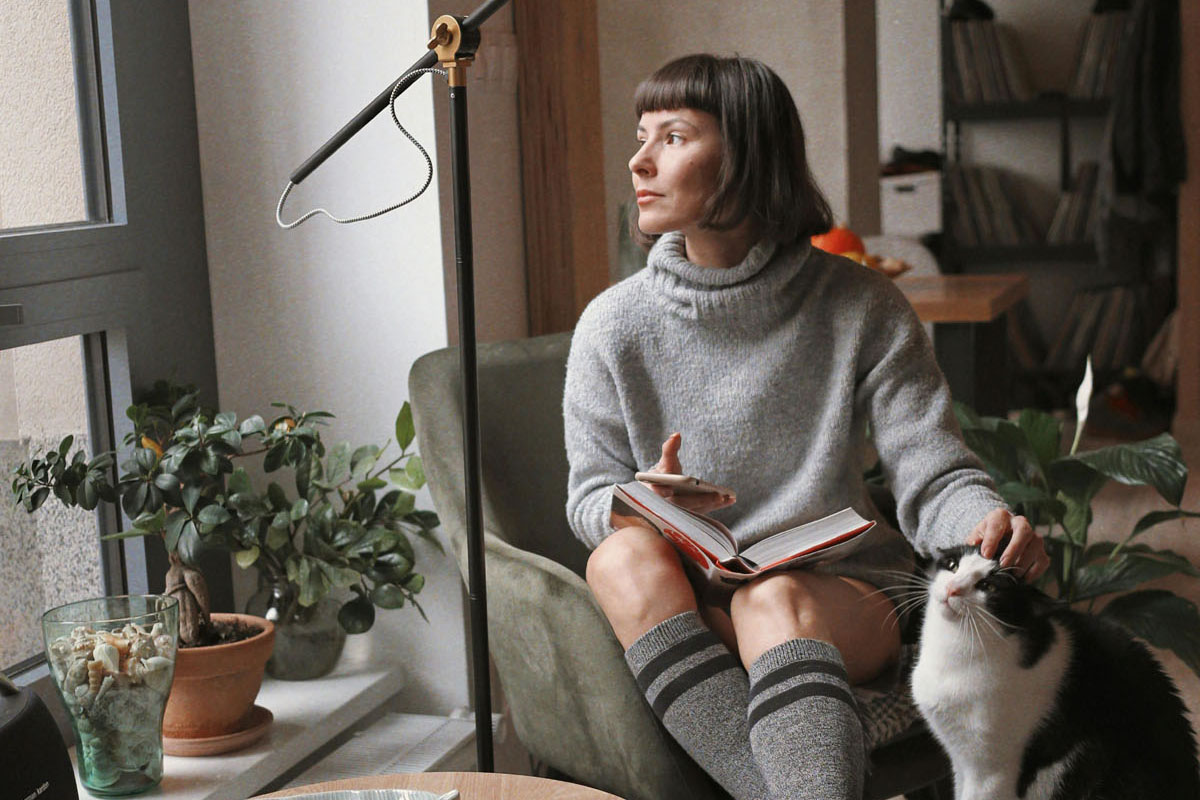 A woman sits petting her cat and looking out the window in the morning. 