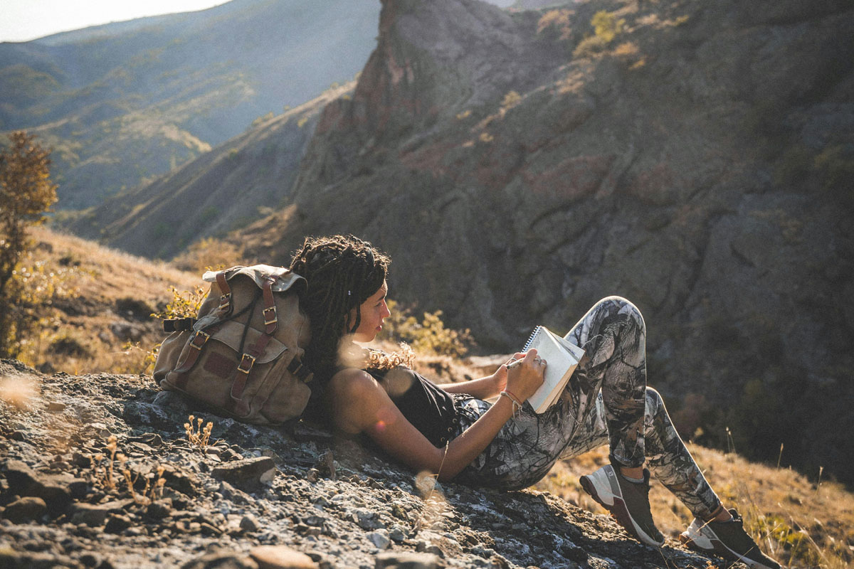 A woman lays on a mountainside writing in a travel journal.