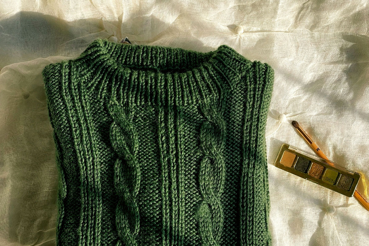 A thick green sweater lays alongside some watercolor paints on a white bed.