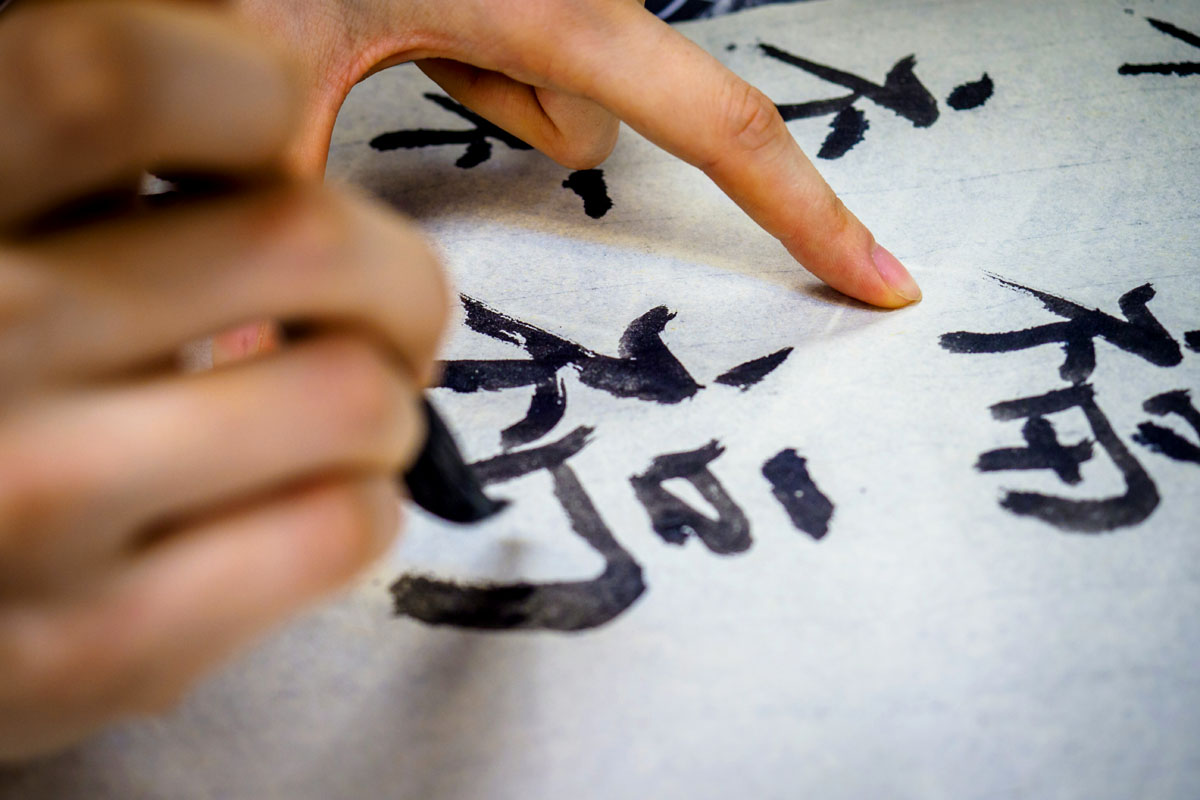 A hand paints Chinese characters in black on white paper.