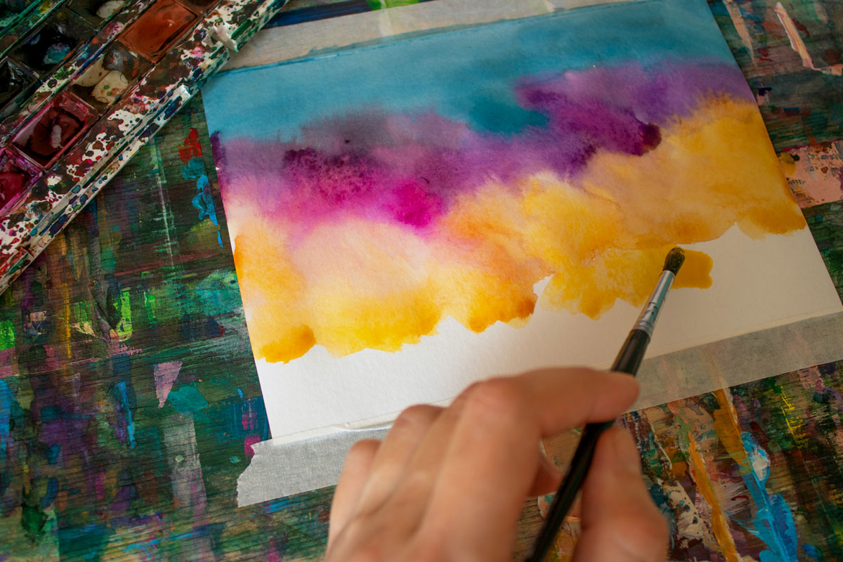 Hands hold a paint brush painting an abstract watercolor with swirls of blue, purple and yellow.