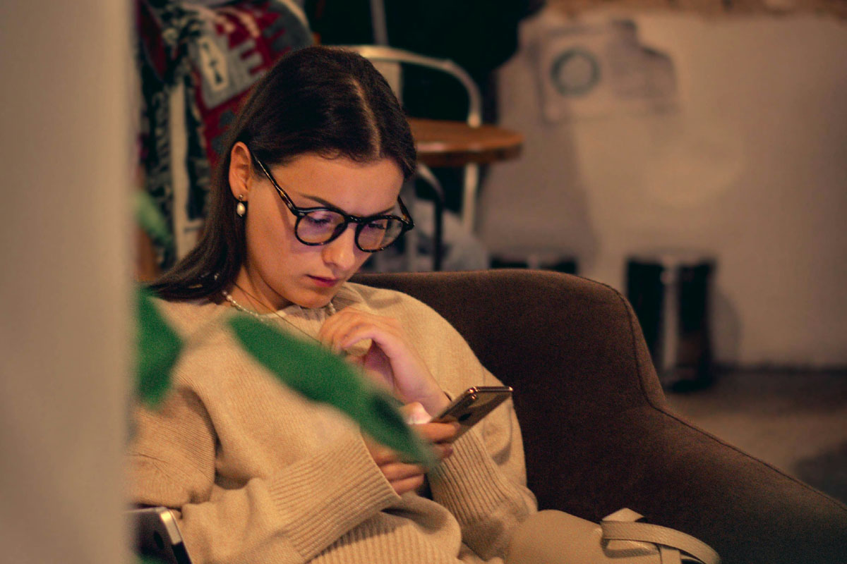 A woman in glasses and a beige sweater sits at a cafe scrolling her phone.