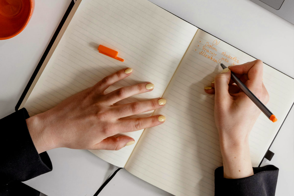 A woman with yellow nail polish writes in an open journal spread on a white table. 