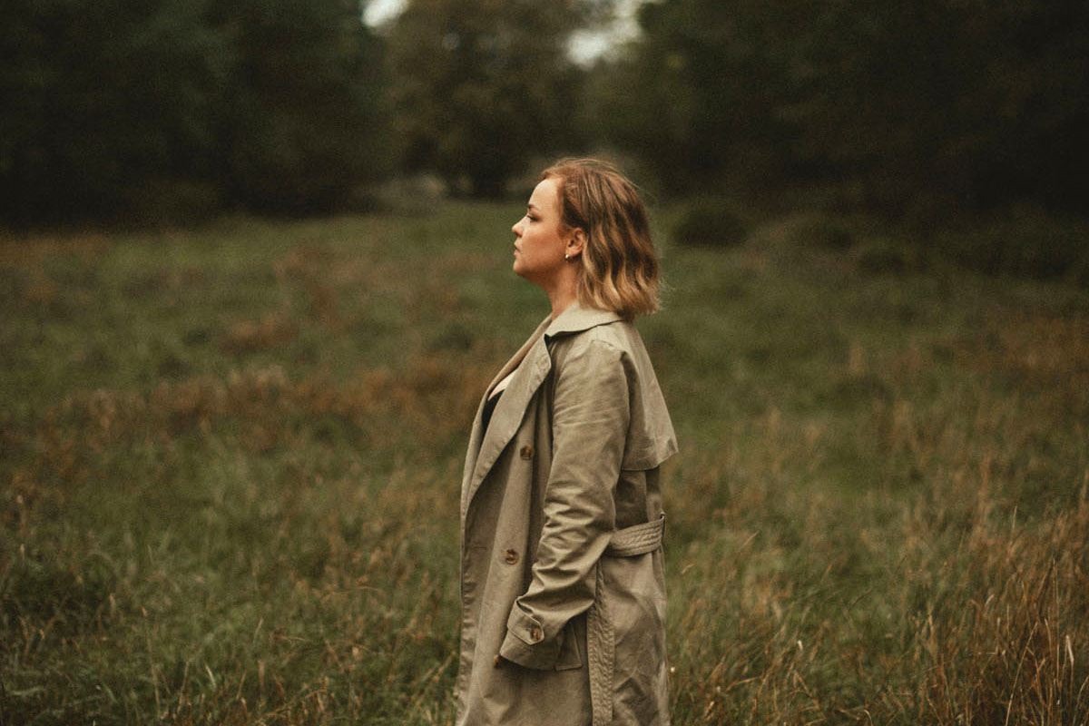A woman in a beige coat stands in a field of grass.