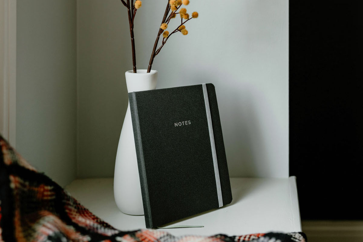 A journal stands on a windowsill next to a white vase. 