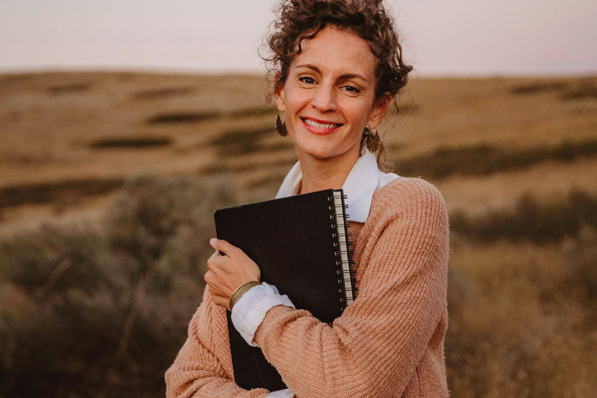 A woman stands in a field holding a black journal. 