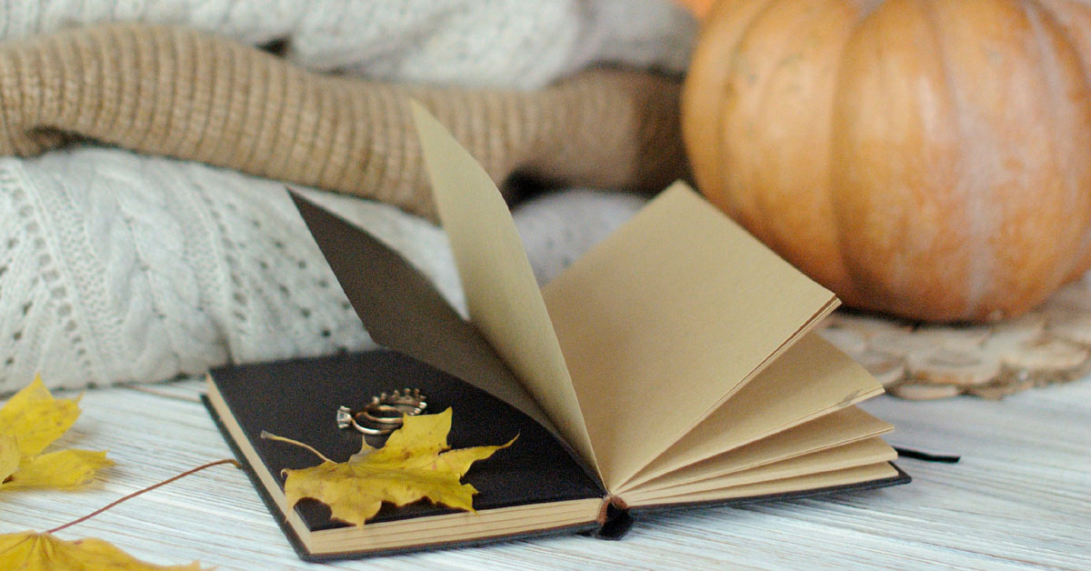 A notebook lays open full of October journal prompts with some leaves, sweaters and a pumpkin alongside.