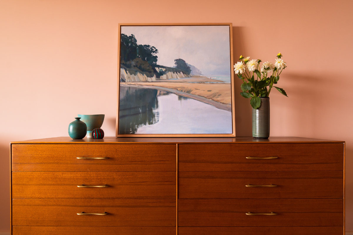 A wooden shelf against a peach wall is lined with vases and a painting. 
