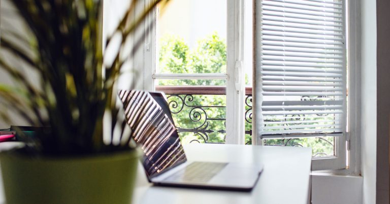 A laptop lays on a white desk with a bright window in the background.