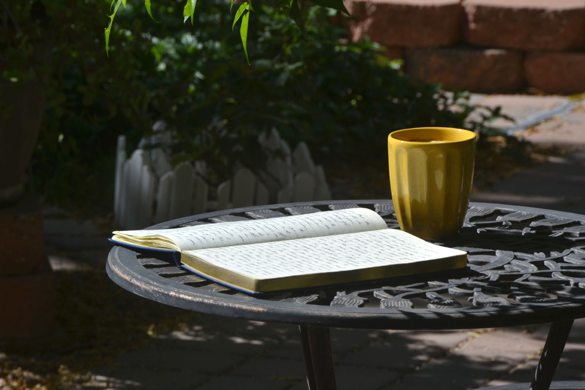 An open paper journal lays on a garden table next to a yellow mug.