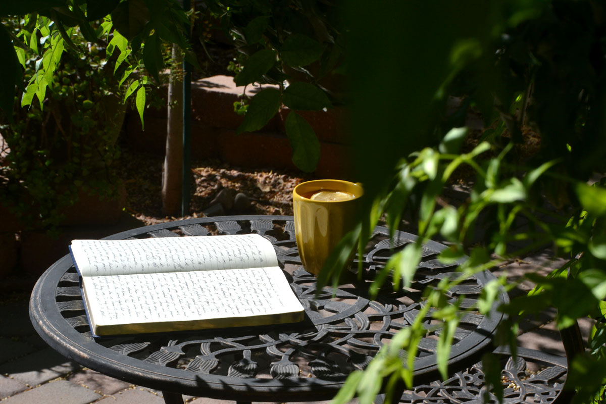 A paper journal lays on a garden table next to greenery and a yellow mug.