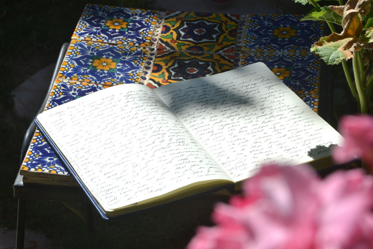 A paper journal lays on a garden table with pink flowers in the foreground.