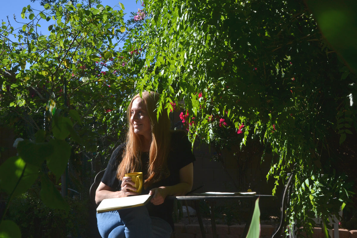 Dee sits in a garden under a large tree smiling and holding a yellow mug in an example of a summer bucket list idea.