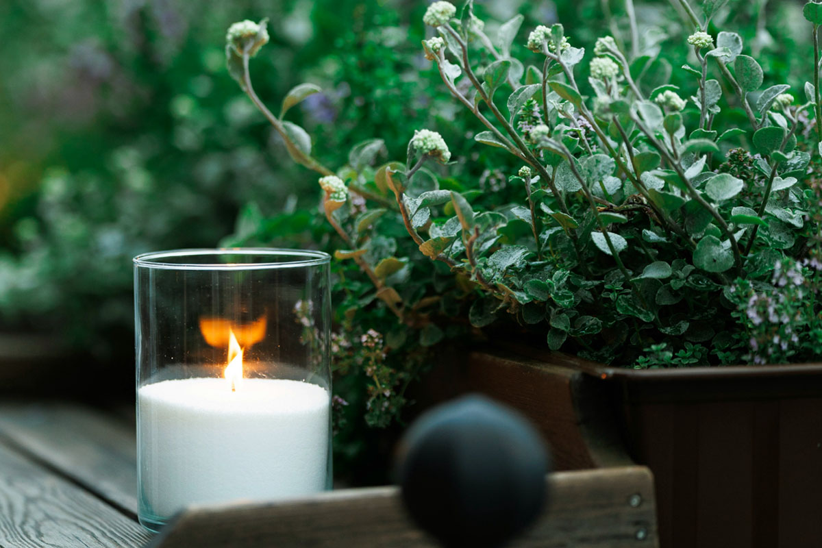A scented candle is lit in a garden against a backdrop of greenery.