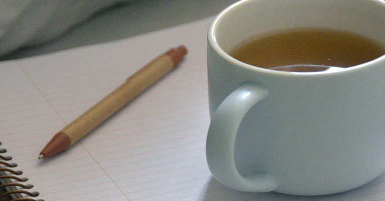 A paper journal lays on a bed next to a mug of tea and a brown pen in an example of an antidote to online journaling apps.