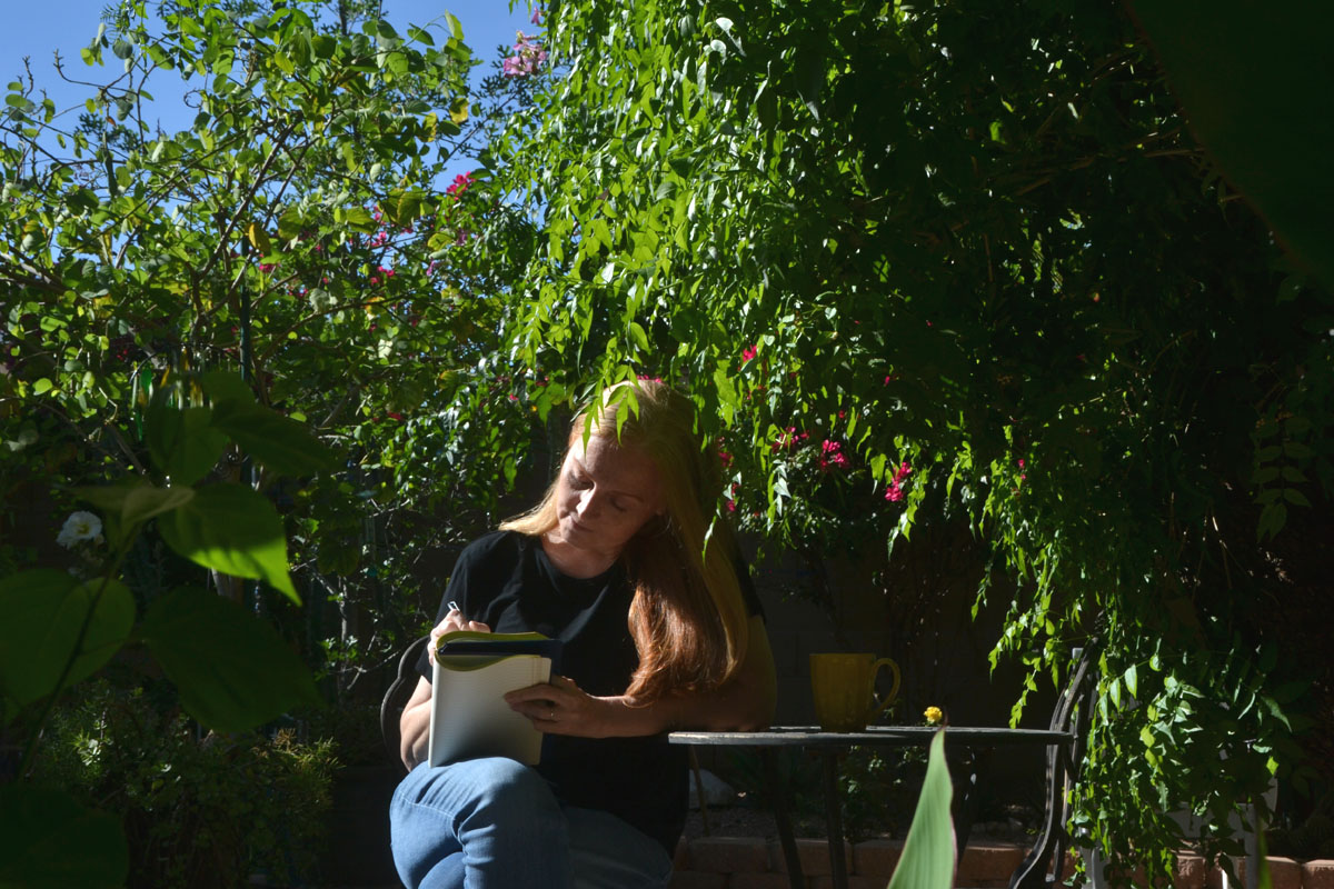 Dee sits in a garden writing with journal prompts over a patio table. 