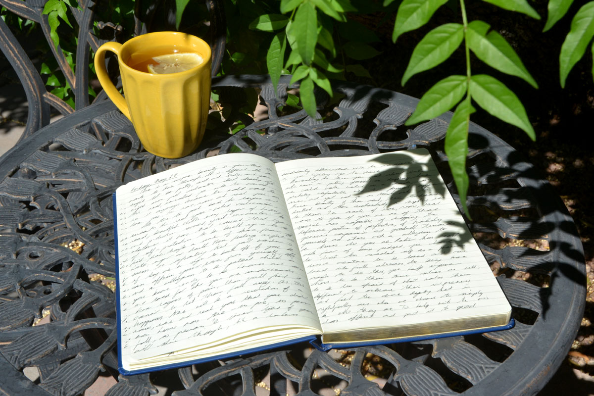 A journal filled with cursive writing lays on a garden table surrounded by greenery.