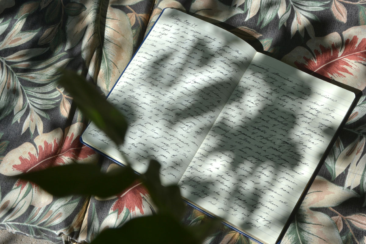 An open journal lays on a floral cushion in a garden.