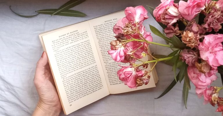An open book lays on a table alongside a vase of pink flowers in an example of reading journal prompts.