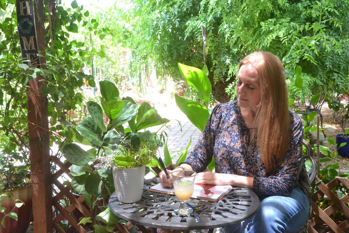 Dee sits in a garden writing in her journal.