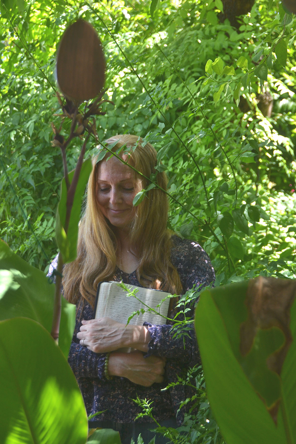 Dee stands in a garden near a tree holding her journal.